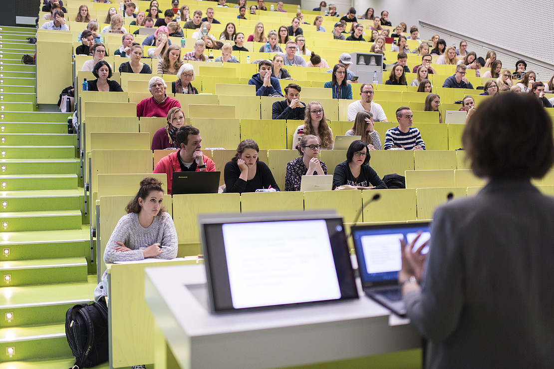 Imagebild: Lehrsituation in einem Hörsaal. Eine Dozentin steht vor ihrem Laptop und spricht zu den Studierenden.